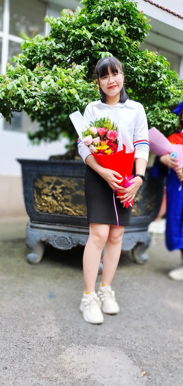 women, adult, plant, full length, smiling, happiness, female, lifestyles, one person, holding, child, architecture, emotion, portrait, young adult, clothing, nature, day, front view, cheerful, outdoors, childhood, potted plant, looking at camera, flower, person, footwear, city, growth, leisure activity, flowering plant, casual clothing, standing, food and drink, spring, business