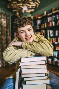 Smiling young man leaning on stack of books while sitting in in bookstore