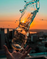 Cropped image of hand catching bottle with splashed water against sky during sunset