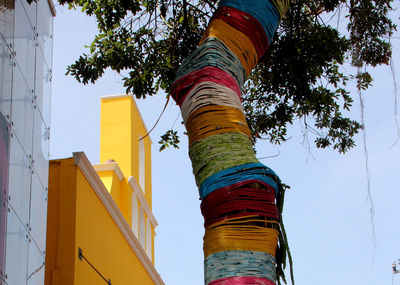 Low angle view of colorful strings tied to tree trunk by buildings