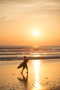 Full length of man with surfboard walking at beach against sky during sunset