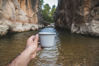 Hands of male tourist standing on the river bank and holding a mug with hot drink.