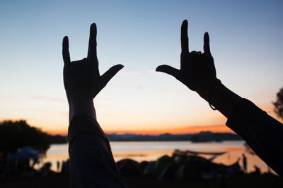 Close-up of silhouette hands gesturing at lakeshore during sunset