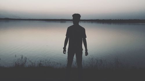 Rear view of silhouette young man standing by lake against sky during sunset