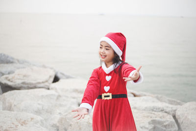 Smiling girl in santa claus costume standing on beach against sea and sky