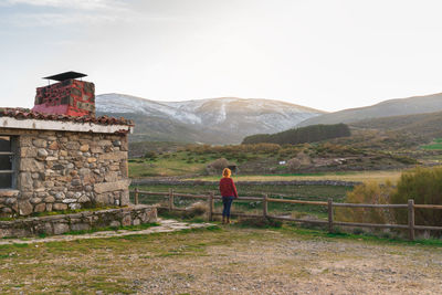 Wide shot of a woman in the doorway of a cabin looking at the landscape with mountains