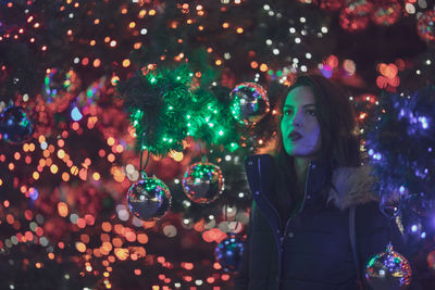 Thoughtful young woman standing amidst illuminated christmas tree at night