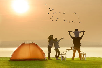 Family enjoying by tent at lakeshore against sky during sunset