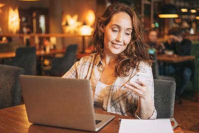 Adult charming brunette woman in plaid shirt working with laptop using mobile phone at cafe