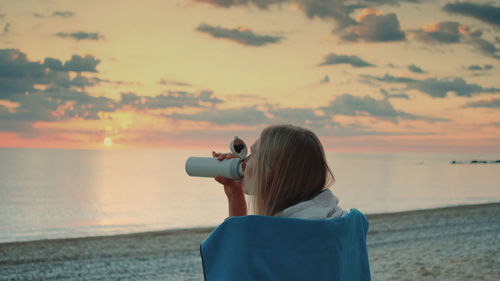 Rear view of woman photographing sea against sky during sunset