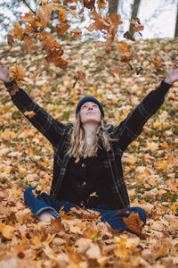 Smiling brunette sitting in a pile of colourful leaves in an oak forest throwing leaves