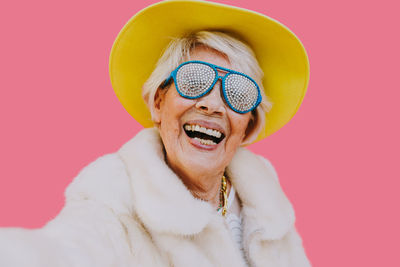 Portrait of cheerful senior woman wearing hat and sunglasses against pink background