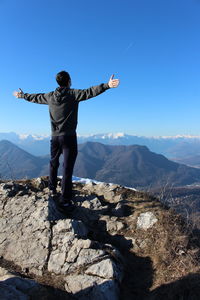 Rear view of hiker standing with arms outstretched standing on cliff by mountains against sky