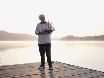 Smiling senior man holding sailing ship while standing on pier