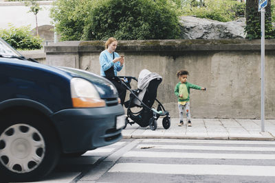 Mother using mobile phone while standing with son and baby stroller on sidewalk at city