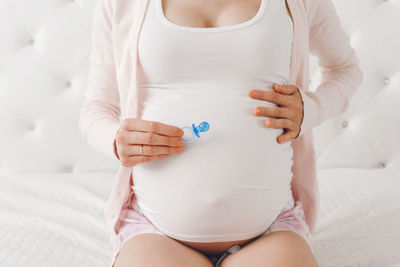 Midsection of pregnant woman holding pacifier sitting on bed at home