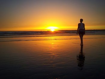 Full length rear view of woman standing at beach against sky during sunset