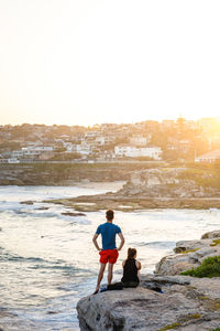 Rear view of couple sitting on rock by sea against sky