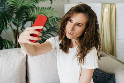 Beautiful woman with wavy hair sits on the sofa at home and takes a selfie on the phone.