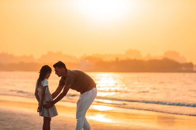 Father and daughter standing on beach against sky during sunset