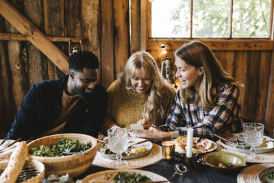 Smiling woman showing smart phone to friends while sitting by dining table during social gathering