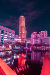 Illuminated modern buildings by river against sky at night