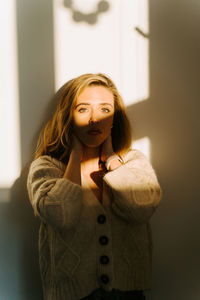Delicate charming female in knitted cardigan standing near wall in room lit by sunlight and looking at camera while touching neck