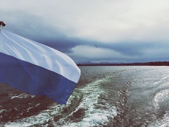 Flag waving above sea against cloudy sky