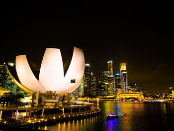 Illuminated modern buildings by river against sky at night