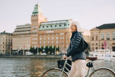 Side view of smiling senior woman with bicycle crossing river in city