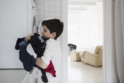 Side view of boy carrying clothes by washing machine at home