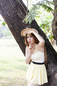 Portrait of beautiful young woman wearing hat while standing by tree trunk