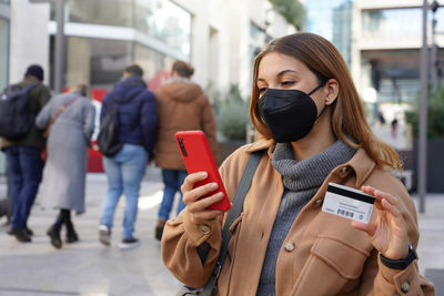 Beautiful young woman with black protective mask ffp2 enters her credit card number on smart phone