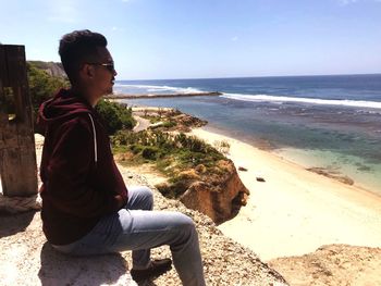 Young man sitting on shore at beach against sky