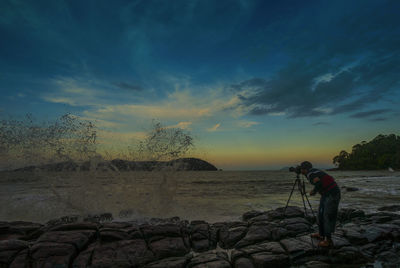 Man photographing sea waves splashing on rocks during sunset