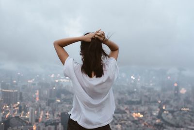 Rear view of woman standing in front of cityscape against sky during foggy weather