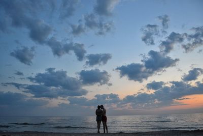 Rear view of men standing at beach against sky during sunset