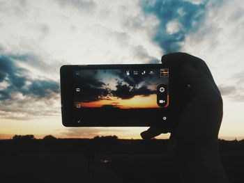 Cropped hand photographing orange sky during sunset in mobile phone