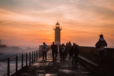 People by lighthouse amidst sea against sky during sunset