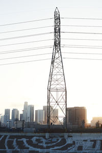 Electricity pylon by buildings against sky during sunset