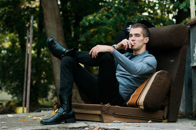 Young man smoking while sitting on sofa against trees