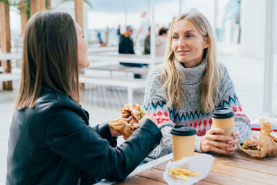 Two girlfriends eat burgers, drink coffee and chat on the street, sitting at the table.