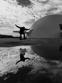 Reflection of man skateboarding in puddle against cloudy sky