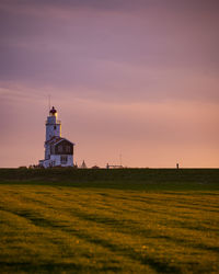 Lighthouse on field by building against sky during sunset