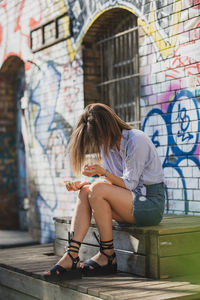 Young woman holding compact disc while sitting on seat against graffiti wall