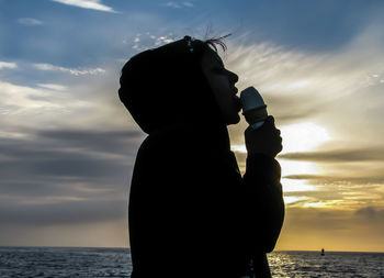 Silhouette boy eating ice cream against sea at sunset