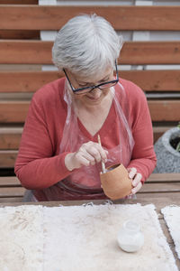 Senior gray haired woman with glasses covering with white glaze handmade clay vase by brush