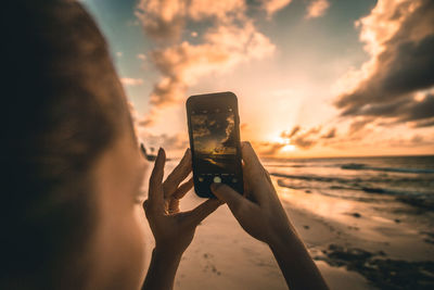 Man photographing through smart phone at beach against sky