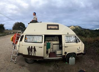 Boy sitting on motor home against cloudy sky