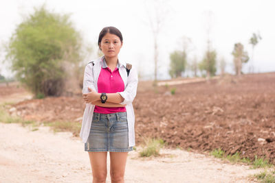 Portrait of young woman with arms crossed standing on field against sky